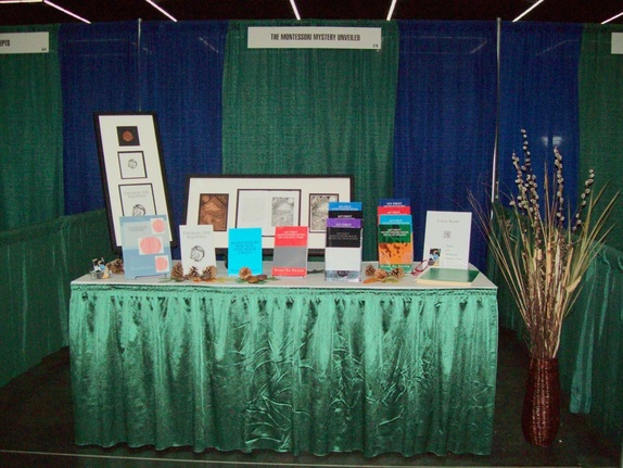 The Montessori Mystery Unveiled vendor booth consists of a solid white surface and green drapes. On top of the table are a dozen different colorful books arranged on single or tiered stands, a shadowbox display of etchings, and tastefully scattered pinecones for decoration. To the right of the display is a wicker vase filled with dried pussy willows and dried grasses.