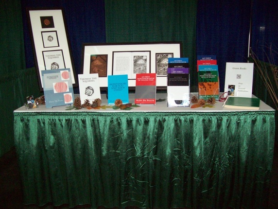 This close-up of the vendor booth showcases the books and etchings. Four books from left to right are propped up by display stands. The eight remaining books are organized into two tiered shelves holding four books each. A notebook and QR code sign are on the far right side of the booth. Etchings of 