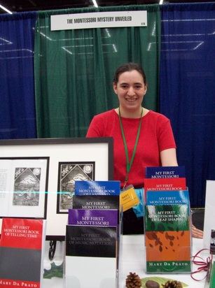 A smiling woman in her early twenties with her dark hair in a ponytail stands behind a vendor booth. Several books are on display in front of her.  A white sign with black letters reading 