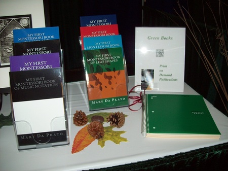 Close-up of far right side of the vendor booth showcases the eight books on two tiered display stands. part of the etching in the background, a green notebook, QR code sign, and a tasteful display of three pinecones on three artificial leaves.